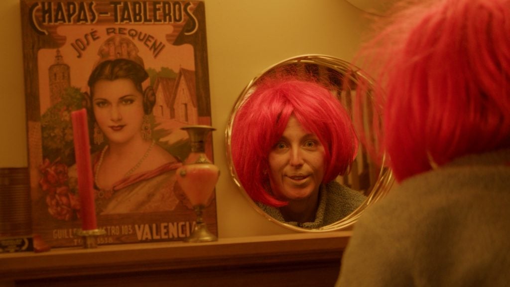 Person with a bright pink wig looks into a round mirror, with a vintage poster of a woman and a candlestick visible on the left side of the image.
