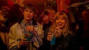 ‘Finding Emily’ Review – A Charming Romance That Finds What’s Been Missing In Rom-Coms Two people holding blue drinks stand in a lively, crowded venue with warm lighting, both looking ahead and smiling as others enjoy the atmosphere around them.