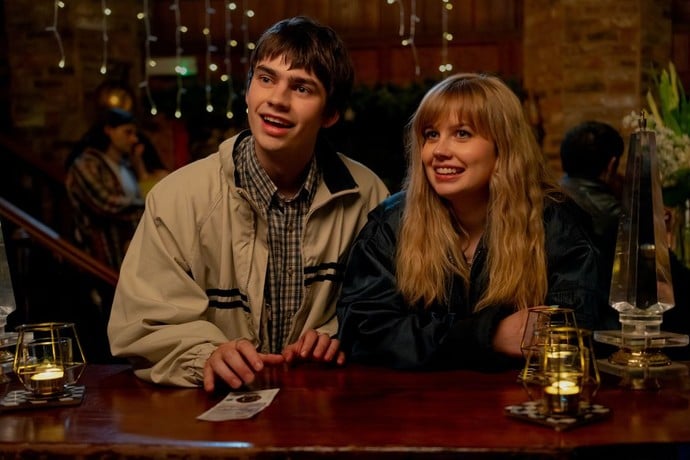 A young man and woman sit at a dimly lit bar, smiling and looking ahead. Small candles and string lights decorate the scene.