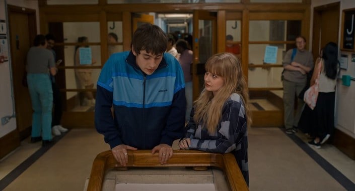 Two people stand at a railing in a hallway, talking. Other people are gathered and walking in the background near doors and bulletin boards.