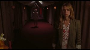 A woman stands in the foreground of a dimly lit hallway while another woman walks away in the background under red lighting.
