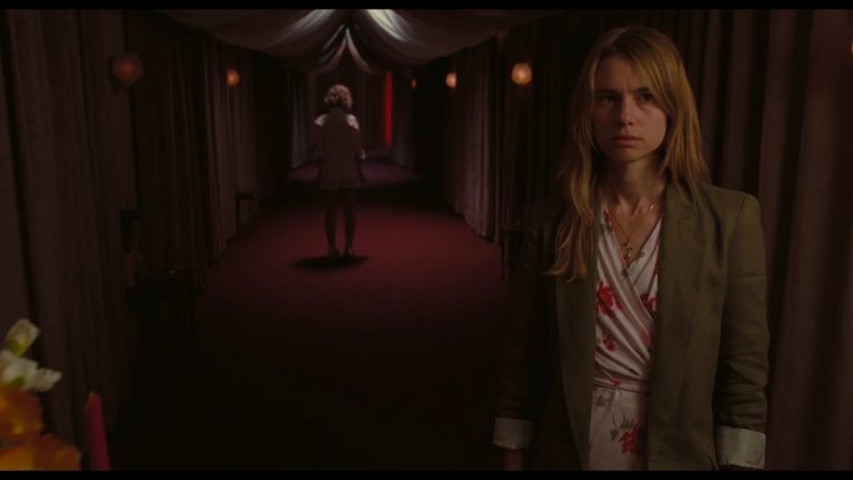 A woman stands in the foreground of a dimly lit hallway while another woman walks away in the background under red lighting.
