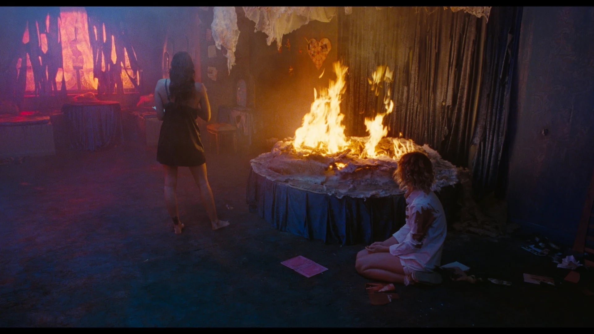 Two women are in a dimly lit room with a large fire burning on a table; one stands facing the fire, and the other kneels on the floor nearby.