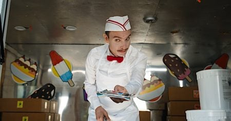 Ice cream vendor in a white uniform and red bow tie stands inside a truck, handing out items, with ice cream decorations on the wall behind him.