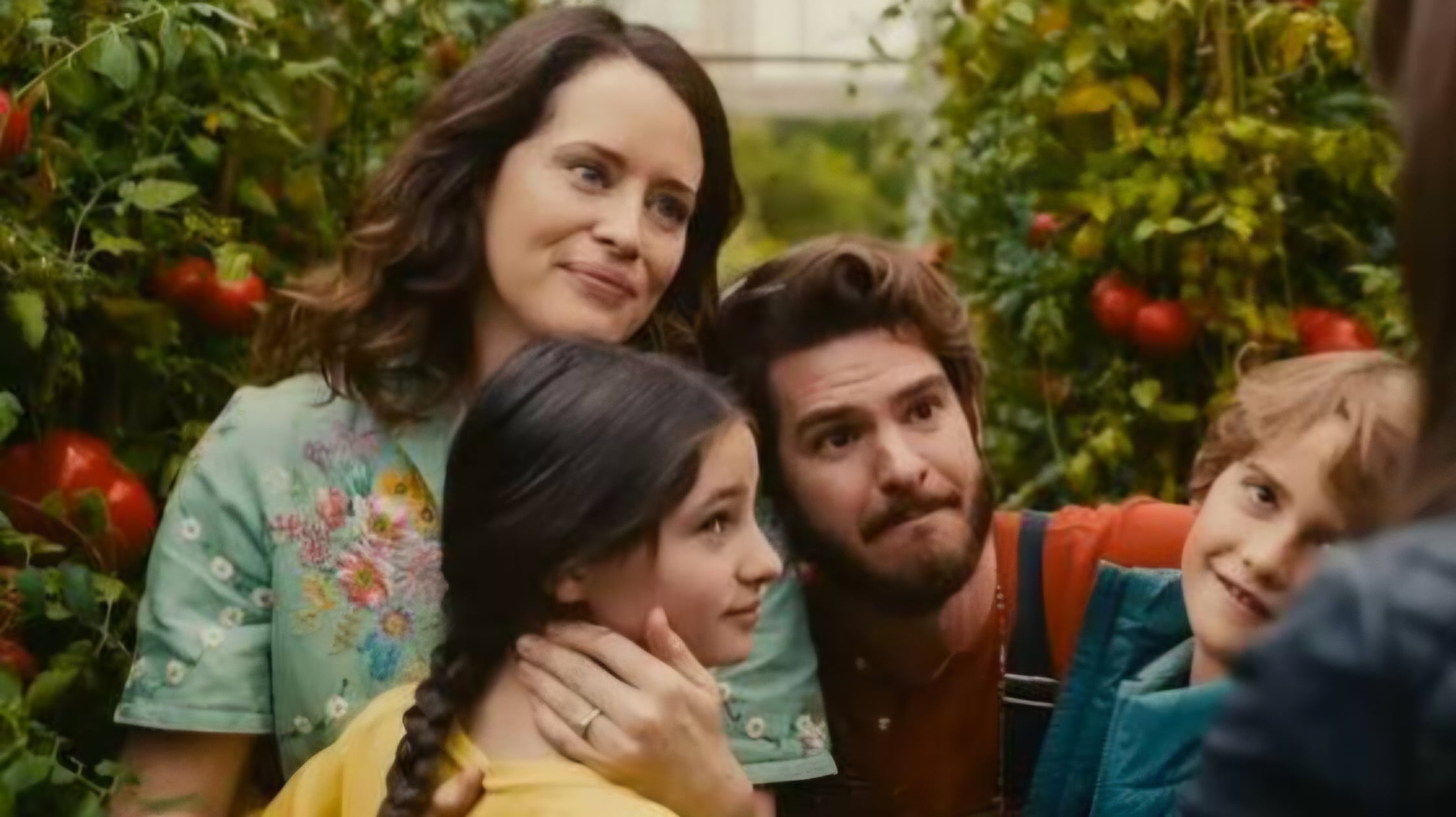 A woman, man, and two children stand closely together in a garden with tomato plants, smiling and looking toward someone outside the frame.