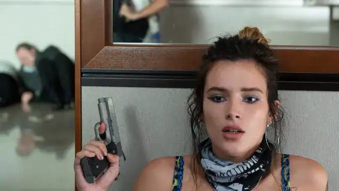 A young woman with a scarf around her neck holds a handgun while sitting against a wall; a blurred figure is visible in the background.
