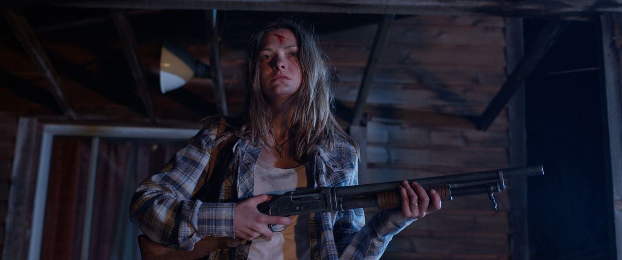 A woman with a bleeding forehead stands under a wooden structure at night, holding a shotgun and wearing a plaid shirt.