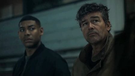 Two men stand side by side in an outdoor setting at night, both looking in different directions with serious expressions.