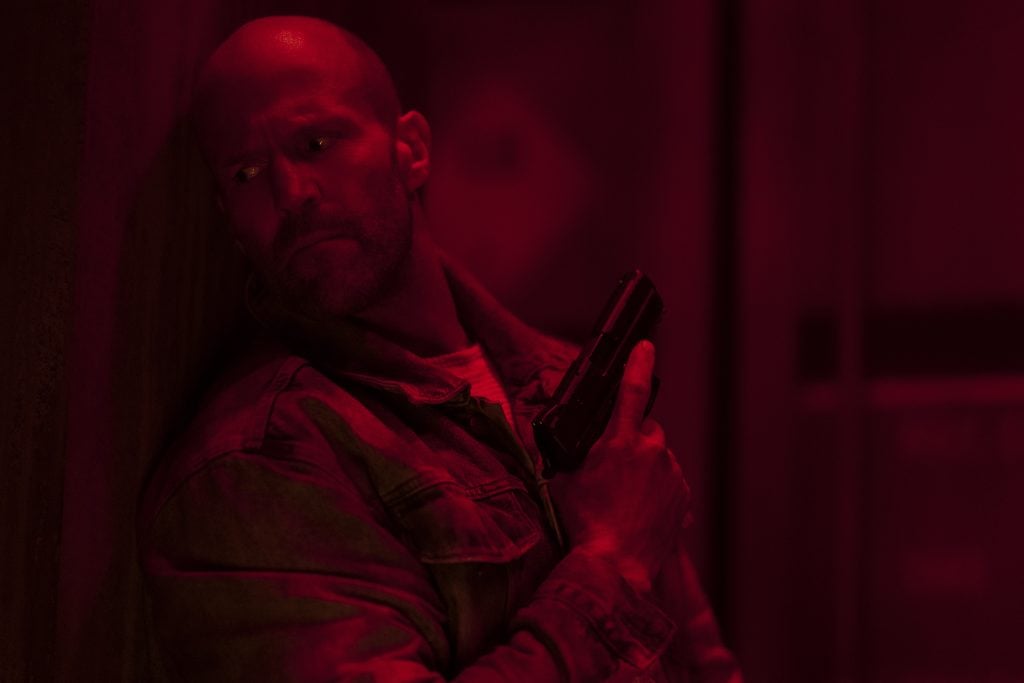 A man with a shaved head, wearing a jacket, holds a handgun and stands against a wall in red lighting, looking to the side.