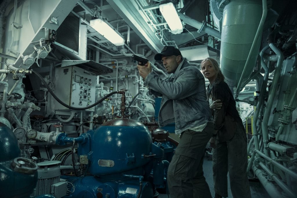 A man holding a gun leads a woman through a cramped, industrial mechanical room filled with pipes, machinery, and overhead lights.