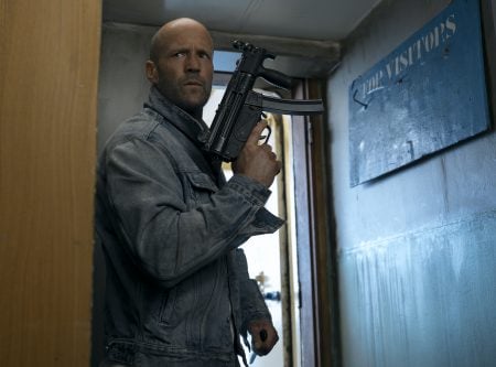 ‘Mutiny’ Review- Another Lean, Mean, Statham Action Machine A man in a denim jacket holds a gun and looks to the side while standing in a doorway next to a blue sign that reads “FOR VISITORS.”.