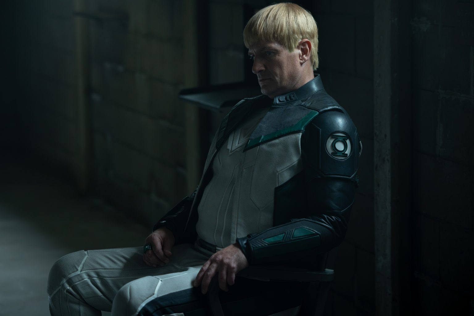 A person in a futuristic suit with green and black details sits on a chair in a dimly lit, industrial room, looking down pensively.