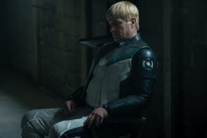 Nathan Fillion’s Guy Gardner Is Quickly Becoming A Key Link Across The New DC Universe A person in a futuristic suit with green and black details sits on a chair in a dimly lit, industrial room, looking down pensively.