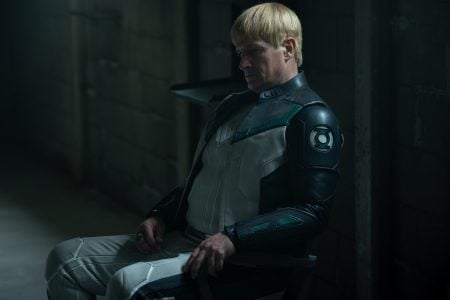 Nathan Fillion’s Guy Gardner Is Quickly Becoming A Key Link Across The New DC Universe A person in a futuristic suit with green and black details sits on a chair in a dimly lit, industrial room, looking down pensively.