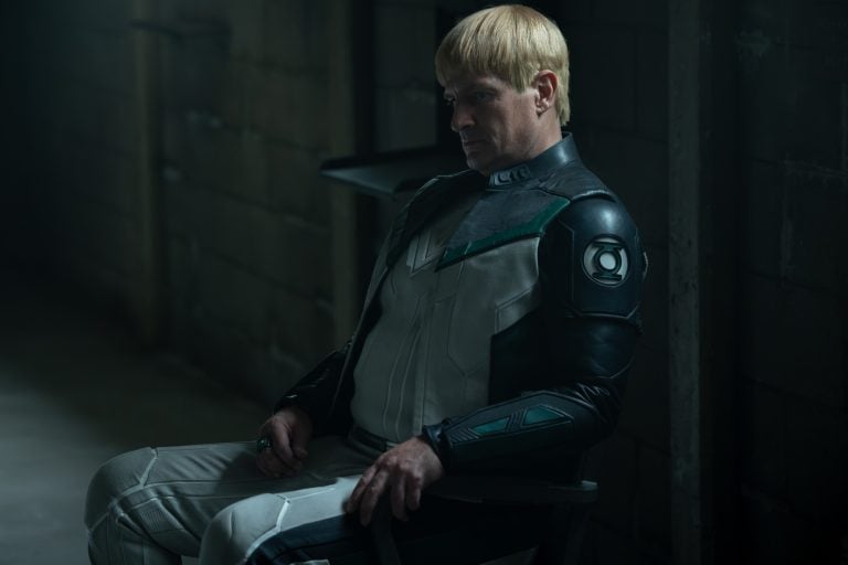 Nathan Fillion’s Guy Gardner Is Quickly Becoming A Key Link Across The New DC Universe A person in a futuristic suit with green and black details sits on a chair in a dimly lit, industrial room, looking down pensively.