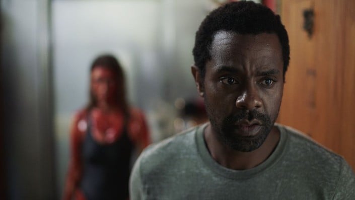 A man in a gray shirt looks worried in the foreground, while a woman covered in blood stands blurred in the background.