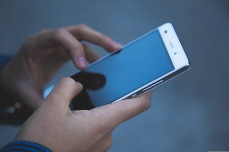 The Modern Fan’s Guide To Understanding Sports Betting Odds A person holds a smartphone with both hands and uses their right thumb to touch the screen. The phone’s screen is off or locked, reflecting light.