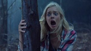 This Canceled Sam Raimi Series Is Suddenly Crushing The Streaming Charts A young woman with long blonde hair and a plaid shirt clings to a tree in a dark forest, looking terrified with wide eyes and mouth open in a scream.