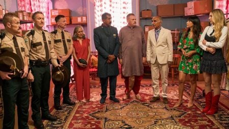 A group of people, including several in sheriff uniforms and others in formal or casual attire, stand in a circle on a patterned rug in a room with shelves and boxes.