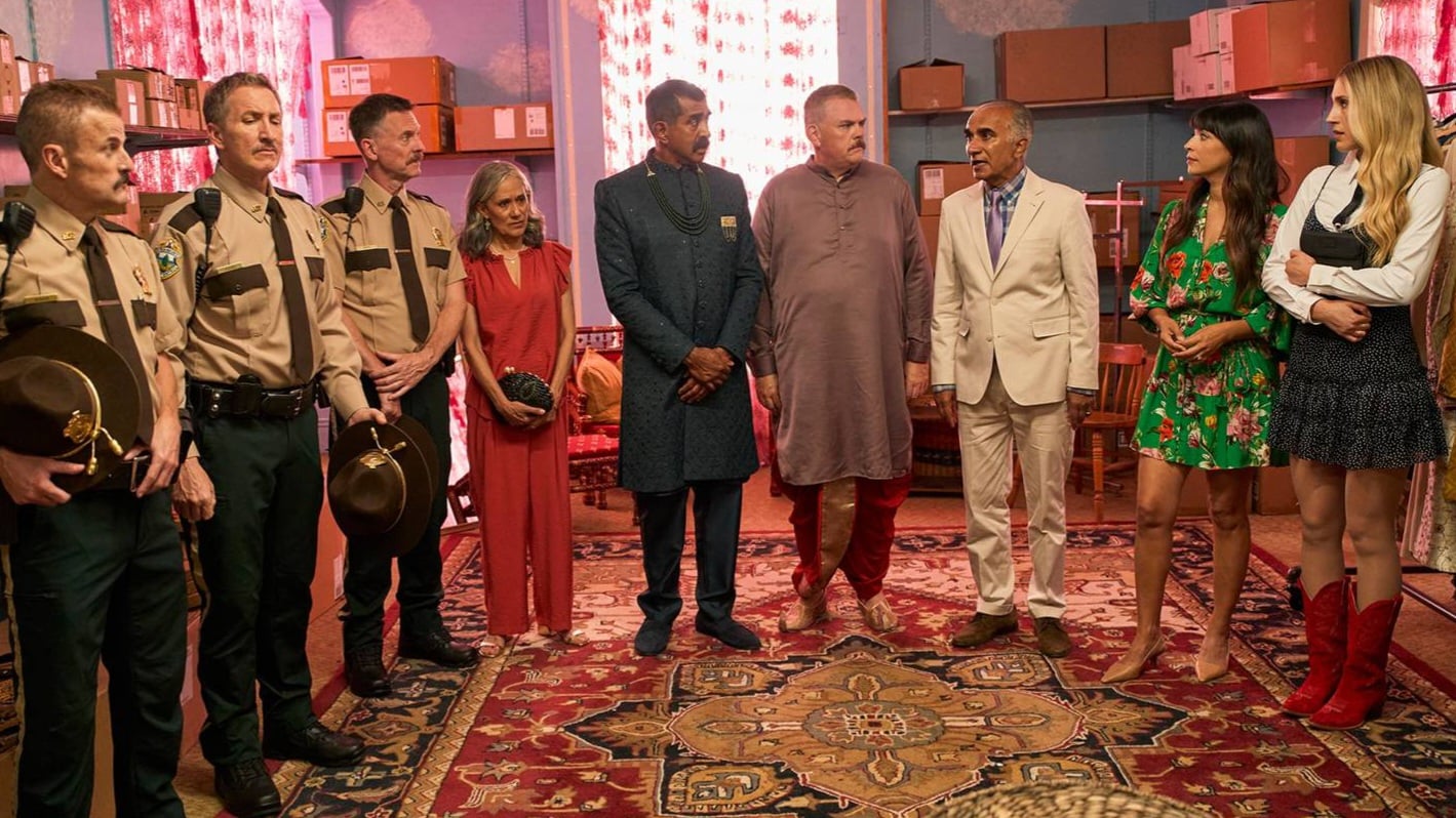 A group of people, including several in sheriff uniforms and others in formal or casual attire, stand in a circle on a patterned rug in a room with shelves and boxes.