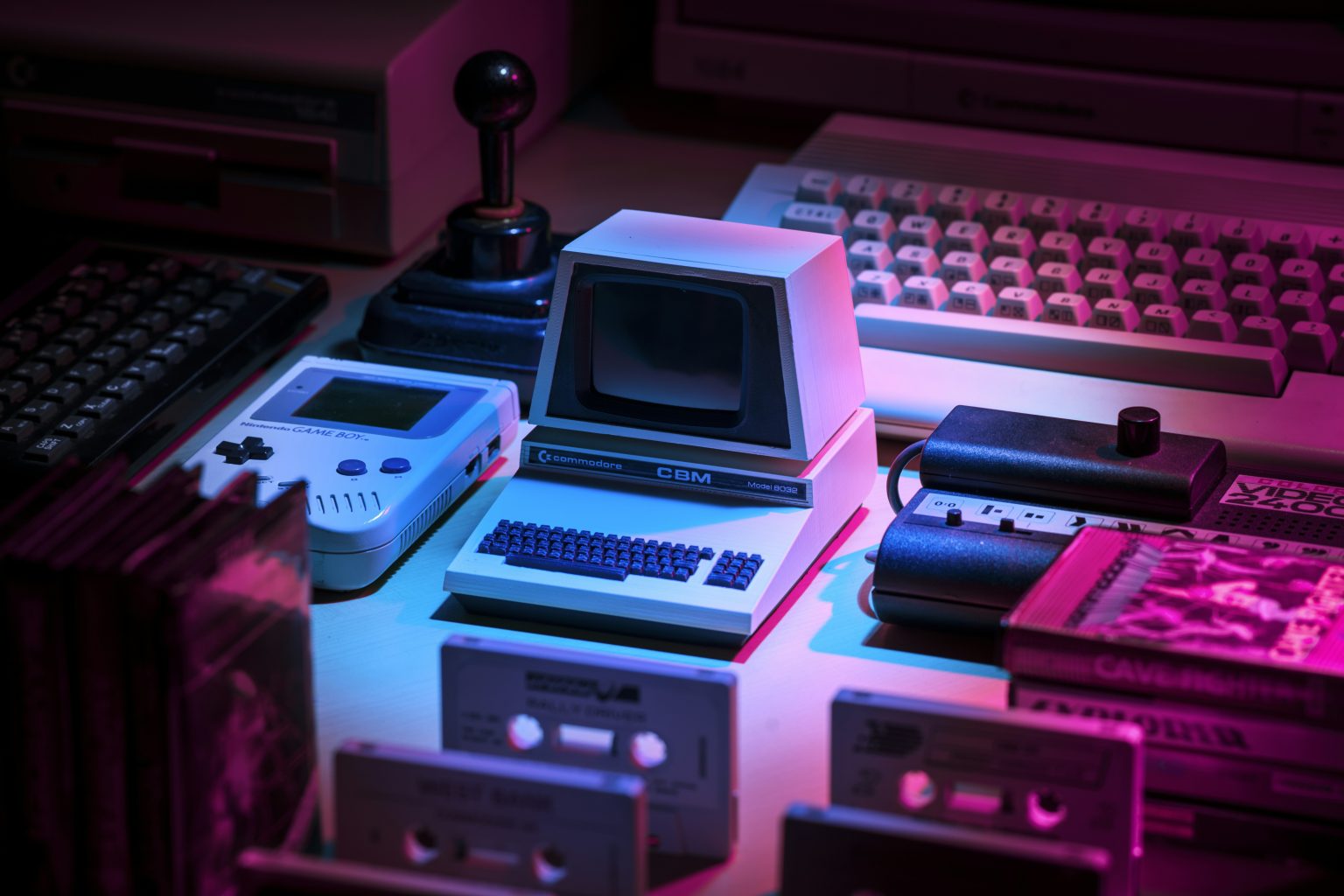 A vintage computer, Game Boy, joystick, keyboard, game cartridges, and cassette tapes are arranged on a table under pink and blue lighting.