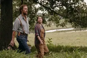 A man and a young girl in overalls walk together through a grassy, rural area with trees and a river in the background.