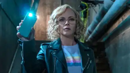 A woman with curly blonde hair and glasses holds up a lit smartphone in a dimly lit industrial setting, wearing a black leather jacket and a pastel-striped shirt.