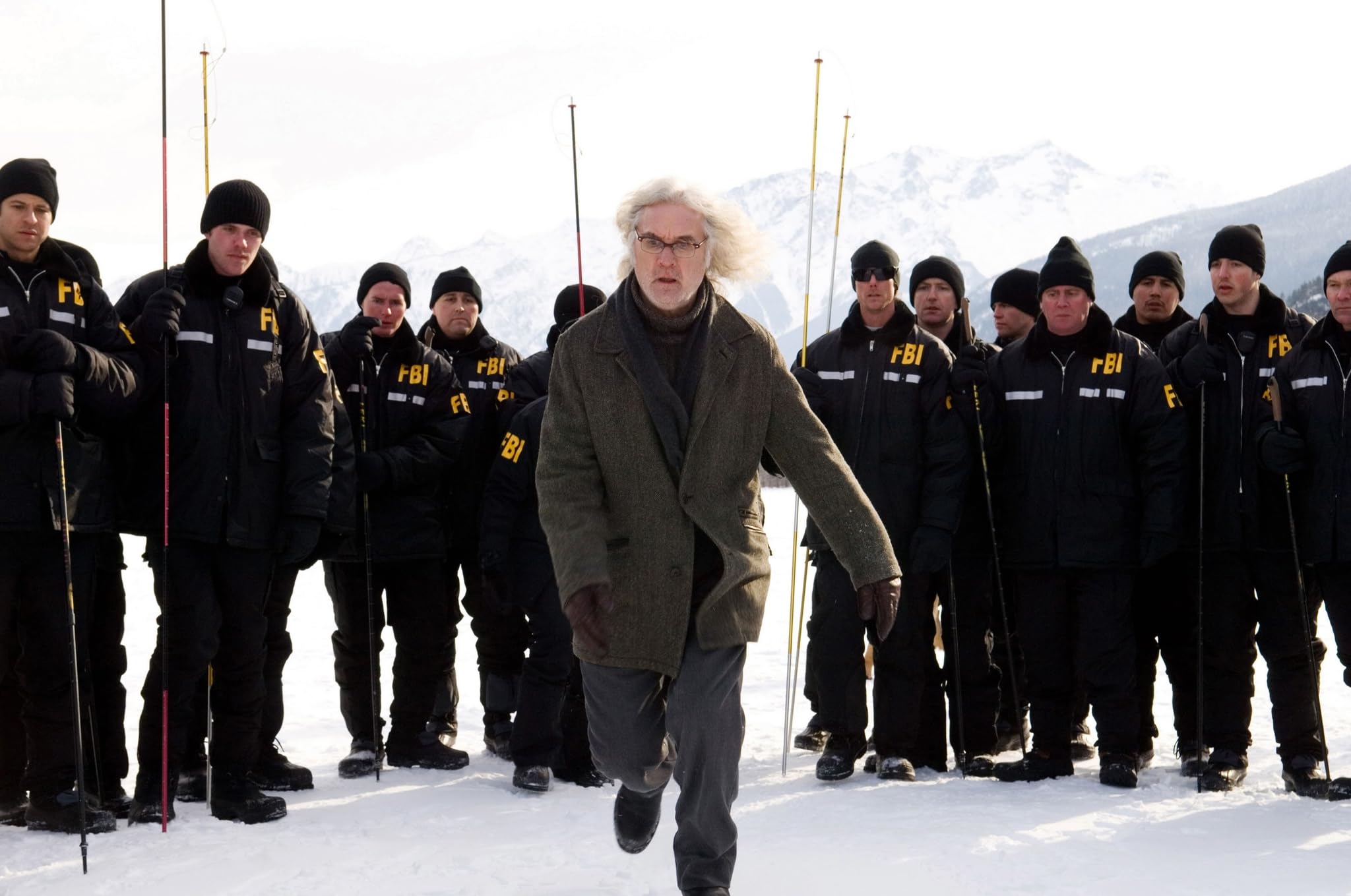 An older man in a coat walks on snow toward the camera, while a group of FBI agents in winter gear stand behind him, holding poles. Snow-covered mountains are visible in the background.