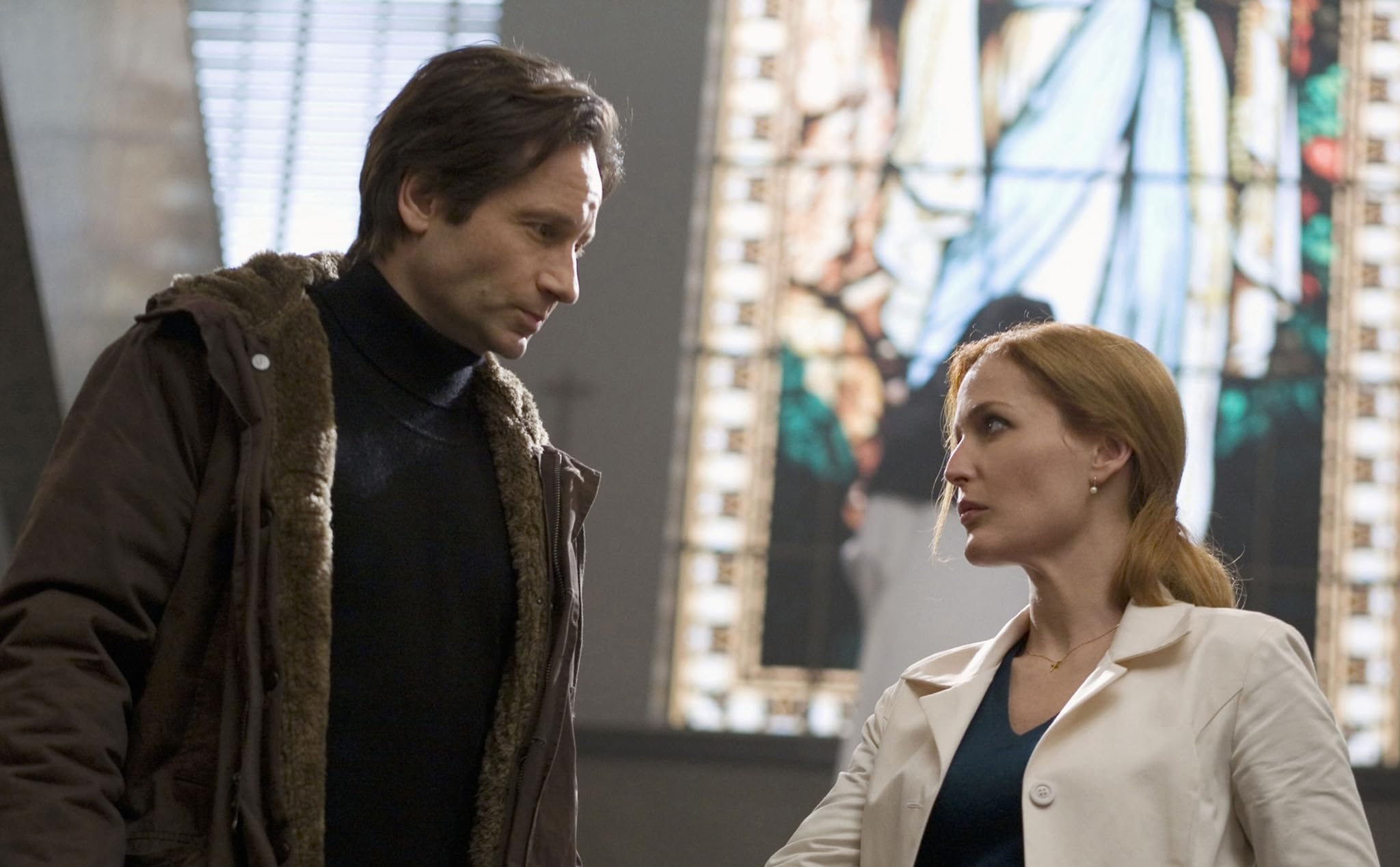 A man in a brown coat and a woman in a white coat stand talking indoors, with a stained glass window visible in the background.
