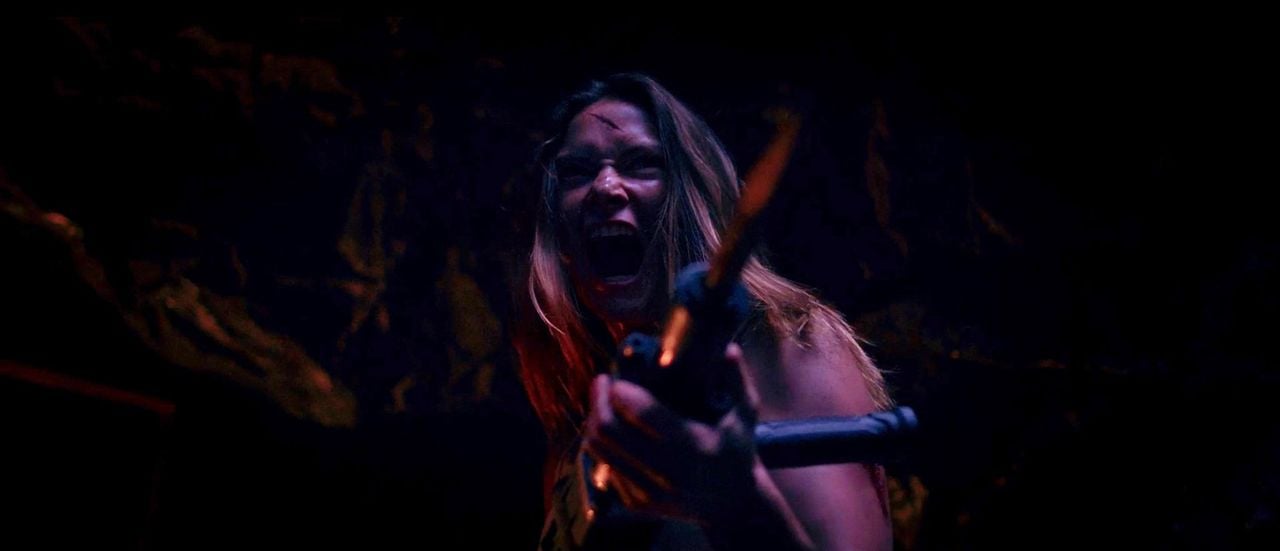 A woman with long hair screams aggressively while holding a weapon in a dimly lit setting with rocky walls.