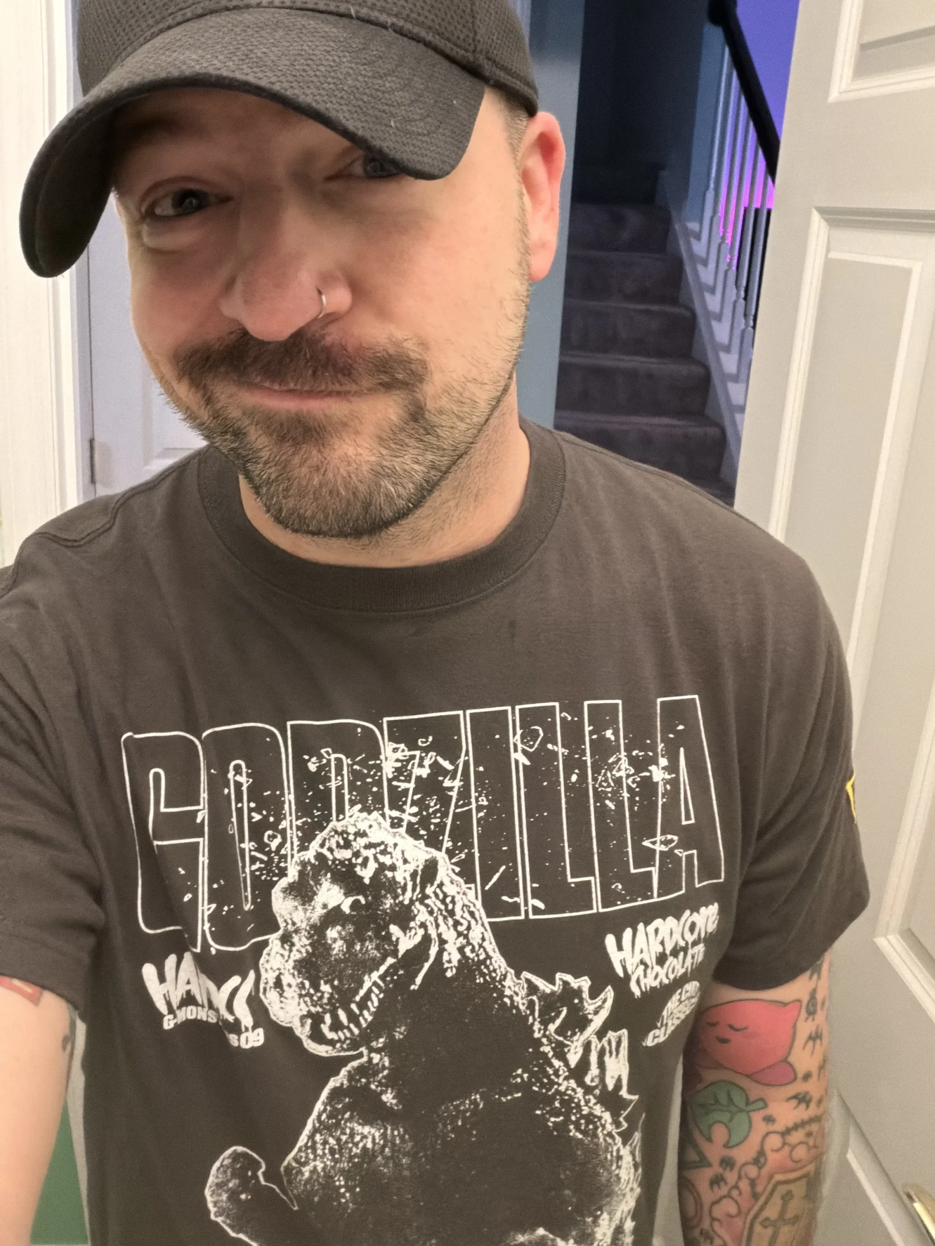 Person with a beard, wearing a black cap and a Godzilla T-shirt, standing indoors near a staircase, smiling slightly.