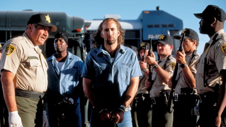 A man in handcuffs stands surrounded by uniformed officers beside a prison transport bus on a sunny day.