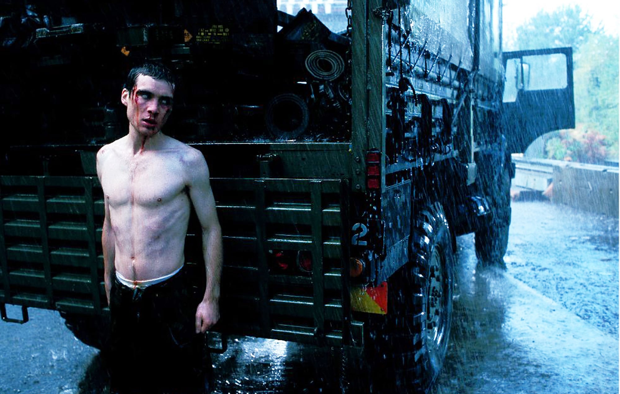 Shirtless man stands in the rain beside a large military-style truck, with wet pavement and trees visible in the background.