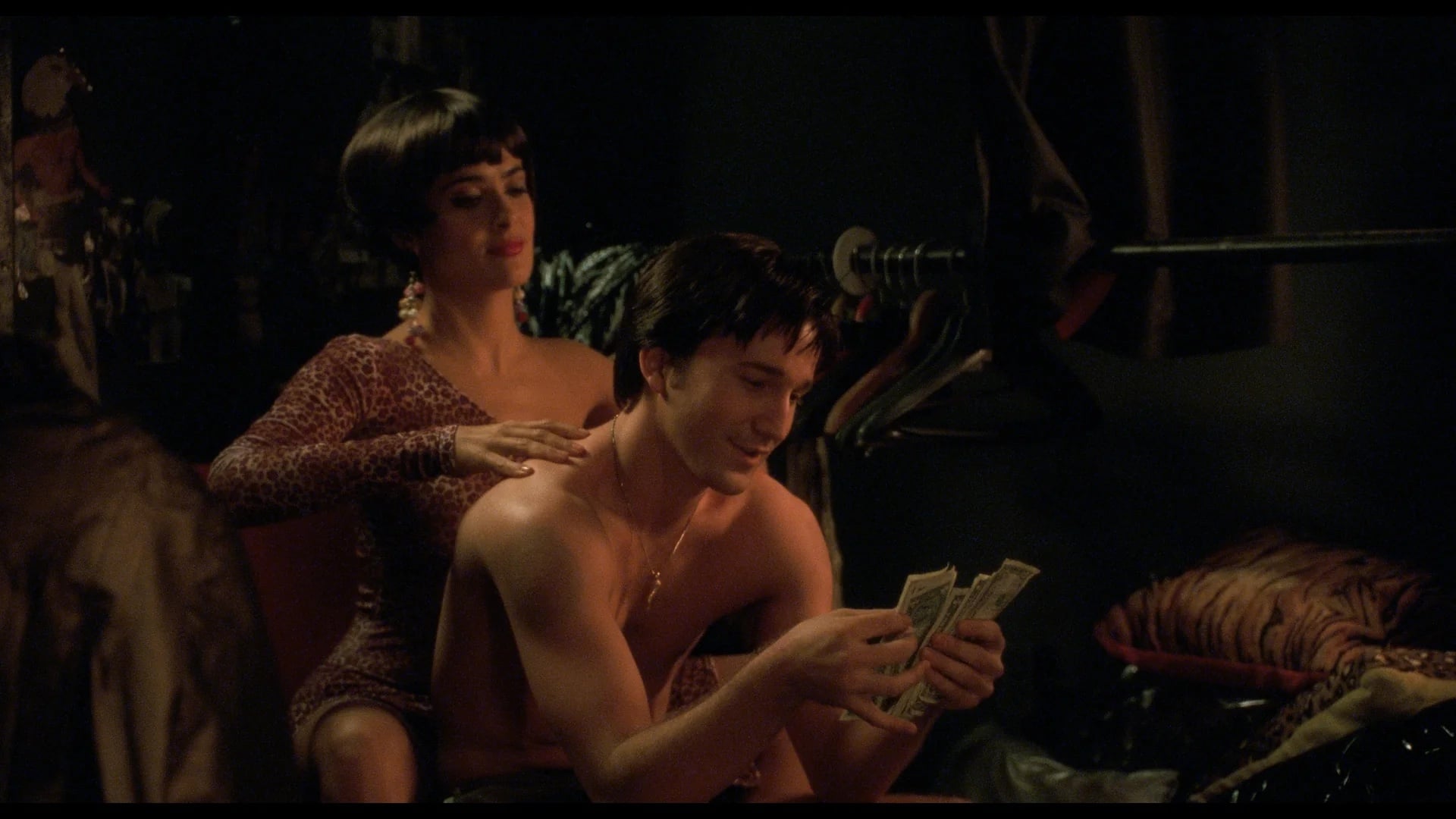 A woman massages a shirtless man who is counting money while sitting in a dimly lit room with clothes hanging in the background.