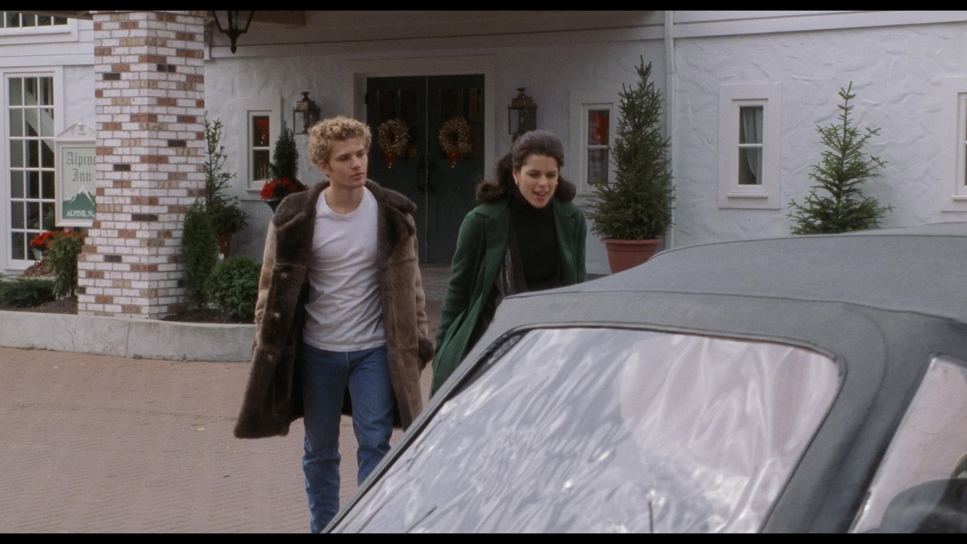 A young man in a fur coat and a woman in a green coat walk toward a parked convertible in front of a building decorated with wreaths and small pine trees.
