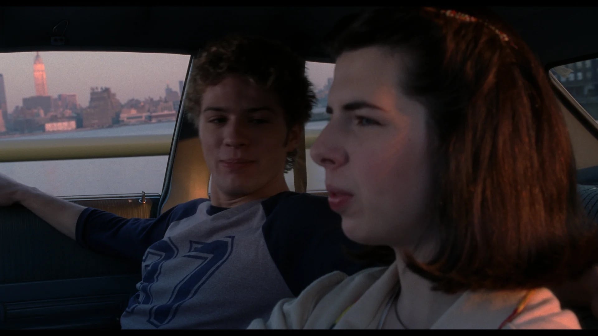 Two young people sit in a car at dusk, with one looking out the window and the other glancing at them. City buildings and a clock tower are visible in the background across the water.