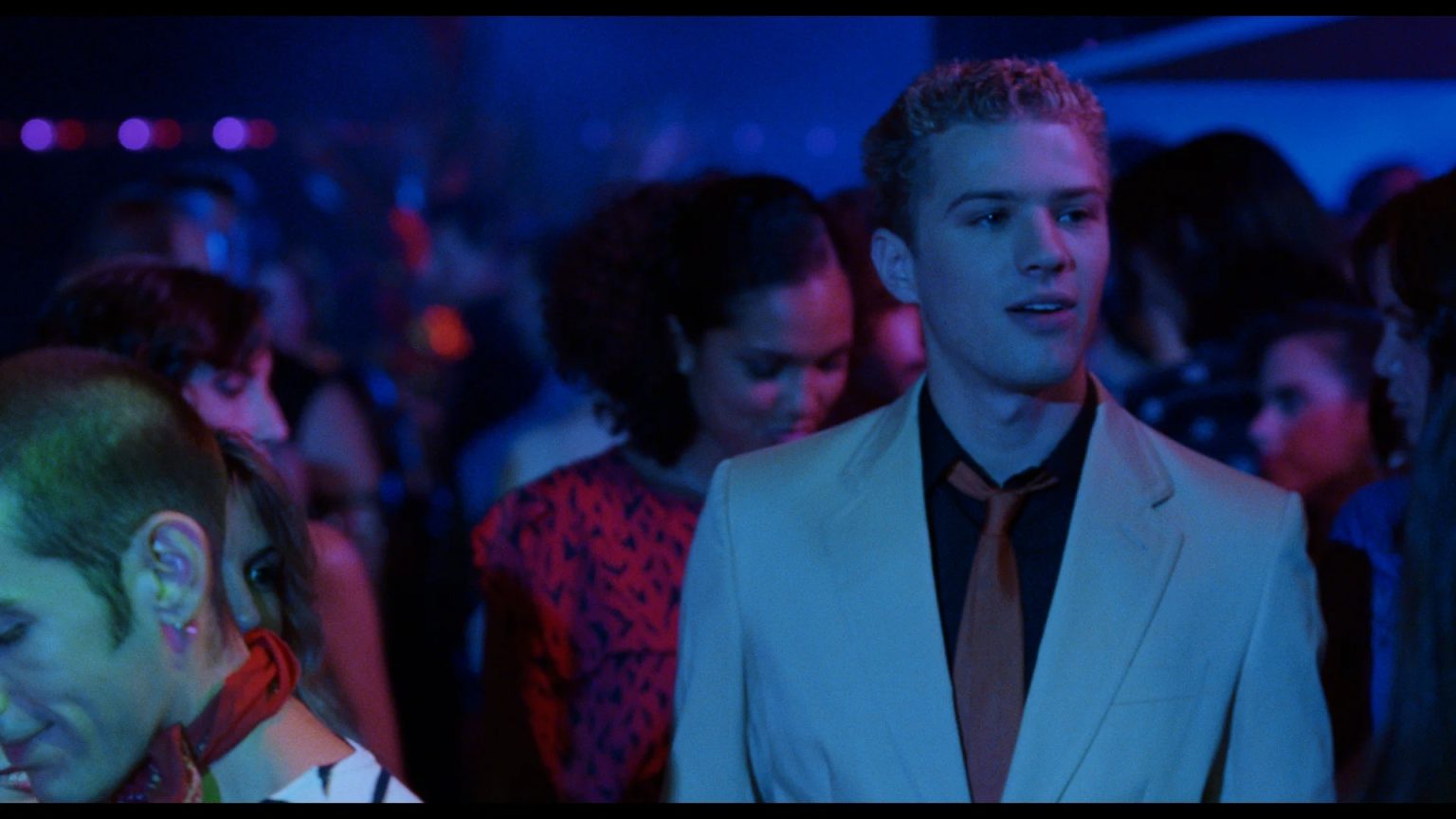 ’54’ 4K UHD Blu-Ray Review – Groovy Period Piece Is A Tale Of Clashing Creative Visions A young man in a beige suit and black shirt stands in a crowded, dimly lit club with colorful lights and people in the background.