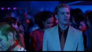 A young man in a beige suit and black shirt stands in a crowded, dimly lit club with colorful lights and people in the background.