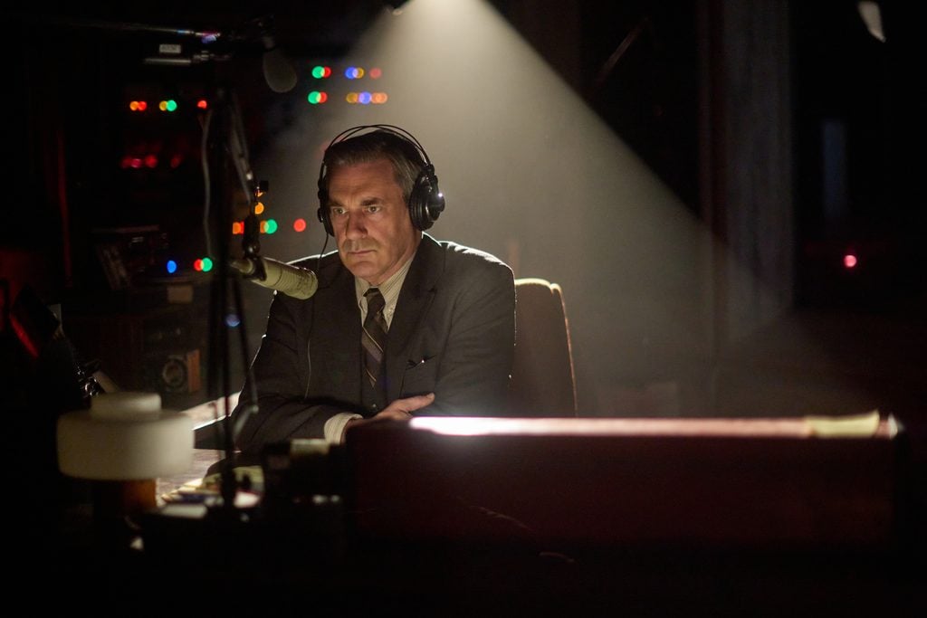 A man in a suit and headphones sits at a desk speaking into a microphone in a dimly lit radio studio with colorful lights in the background.
