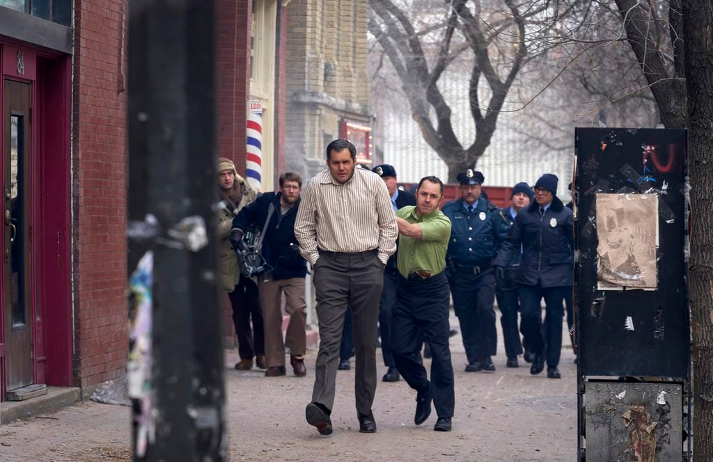 A man in a striped shirt is handcuffed and escorted by another man while police officers and bystanders follow on a city sidewalk.