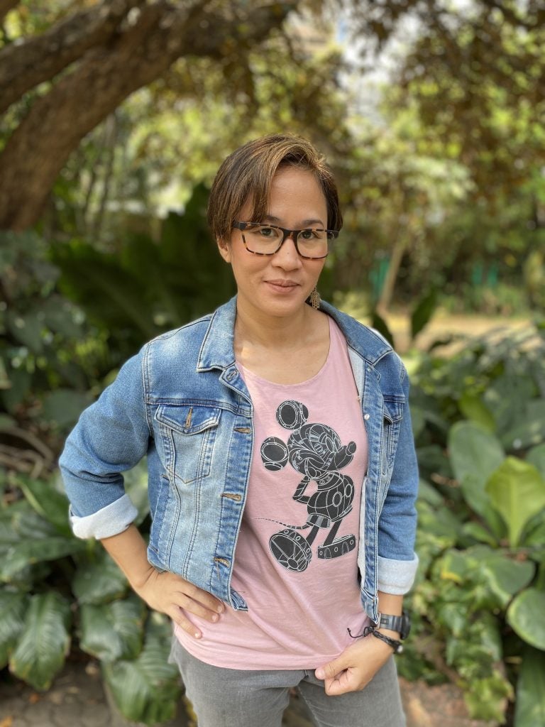 Person with short hair and glasses stands outdoors, wearing a denim jacket over a pink T-shirt with a graphic design, surrounded by green leafy plants and trees.