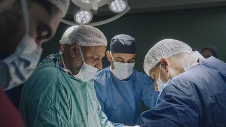 A group of surgeons in scrubs, masks, and hair covers perform surgery under bright operating room lights.