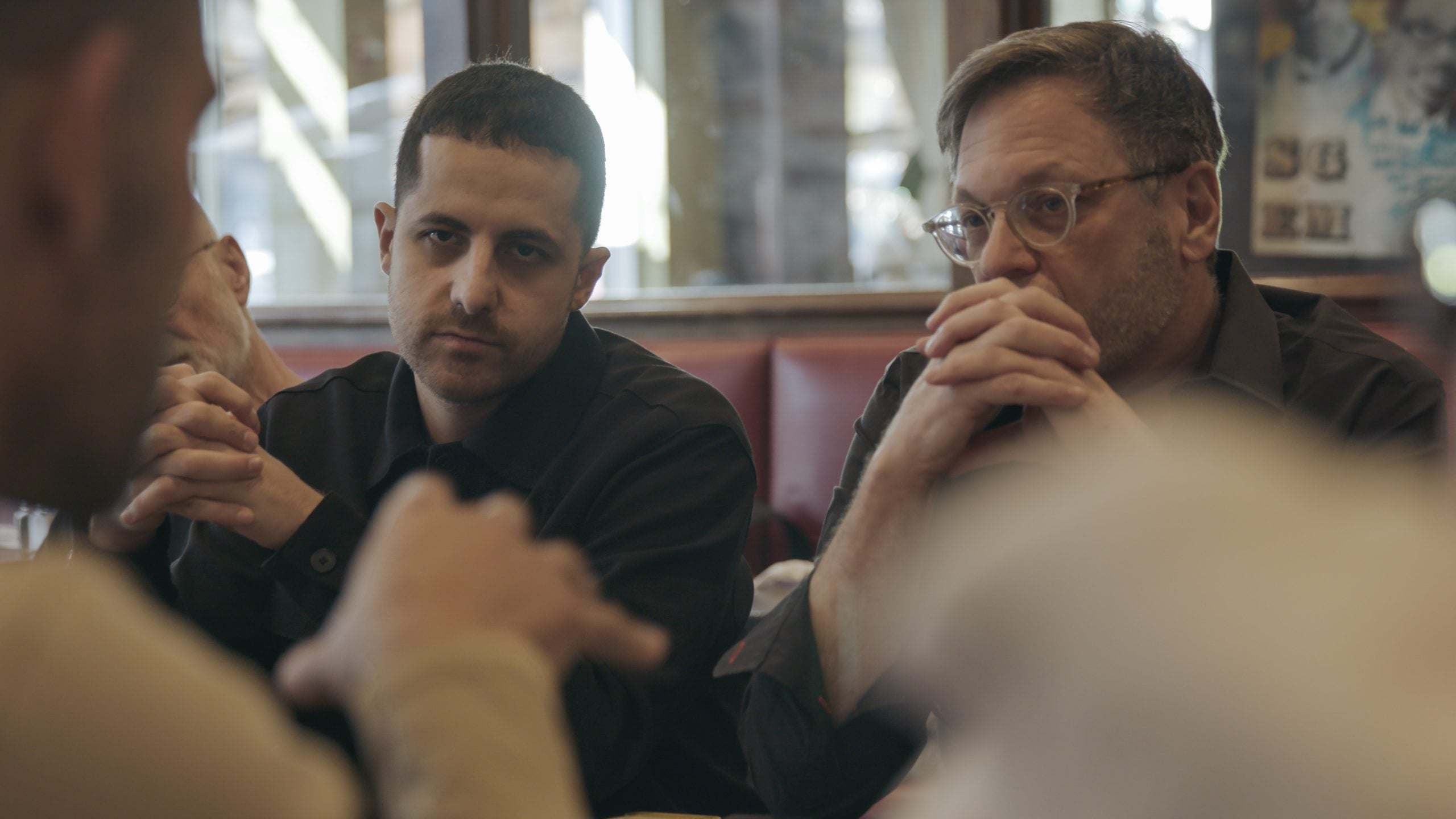 Two men sit at a table in a restaurant, listening intently to someone speaking off-camera.
