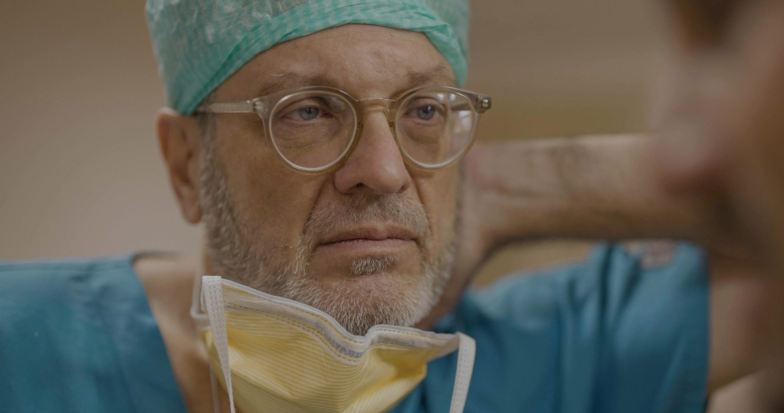 A middle-aged man in scrubs, surgical cap, and glasses lowers his face mask and looks serious, with his hand behind his neck.