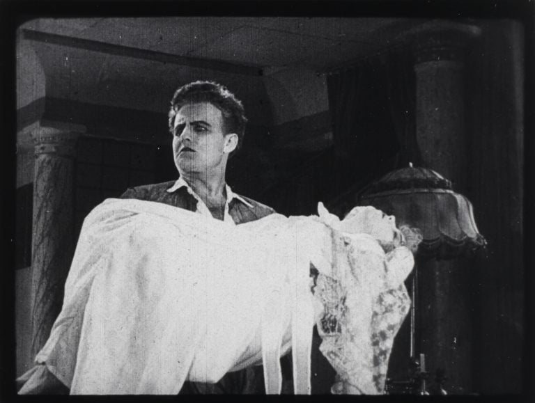 Czech Silent Genre Rarities From Director Jan S. Kolár Blu-Ray Review – A Genre-Bending Duo From The Silent Era A man in period clothing holds an unconscious or lifeless woman in his arms inside an ornate room with columns and a lamp.