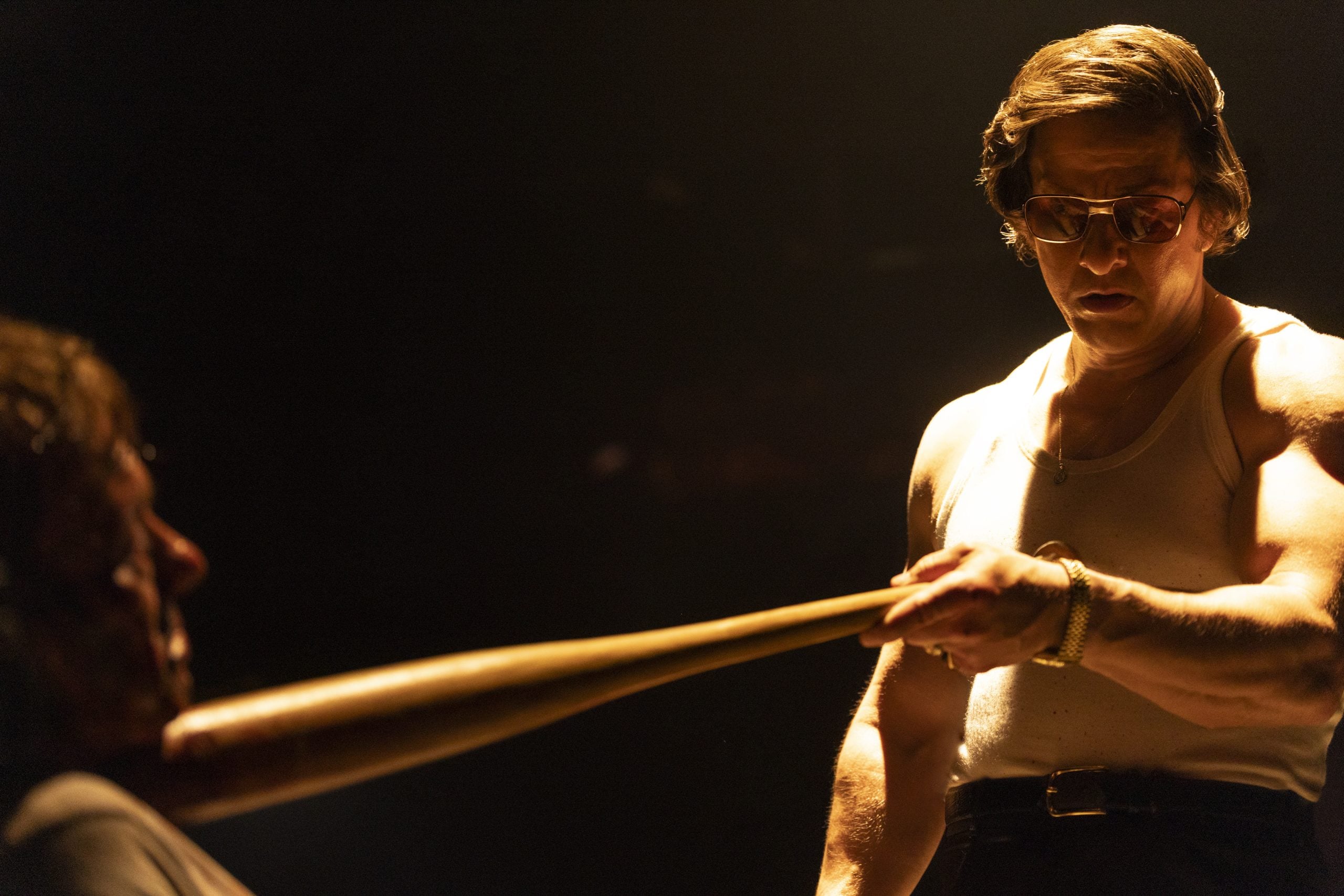 A man in a white tank top holds a baseball bat close to another man’s face under dramatic lighting.