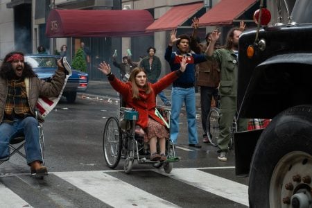 A group of people, including a woman in a wheelchair, raise their hands in front of a large vehicle on a city street during what appears to be a protest.