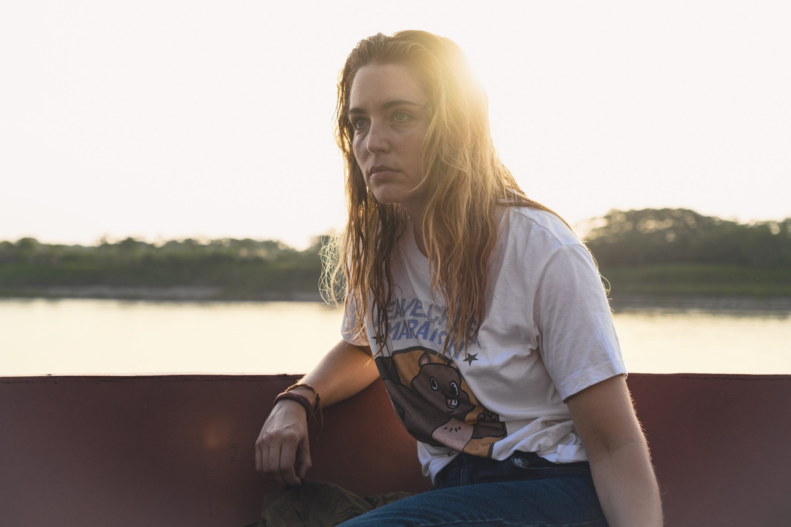 A woman with wet hair sits on a boat at sunset, wearing a white graphic t-shirt and jeans, with the sun behind her and water in the background.