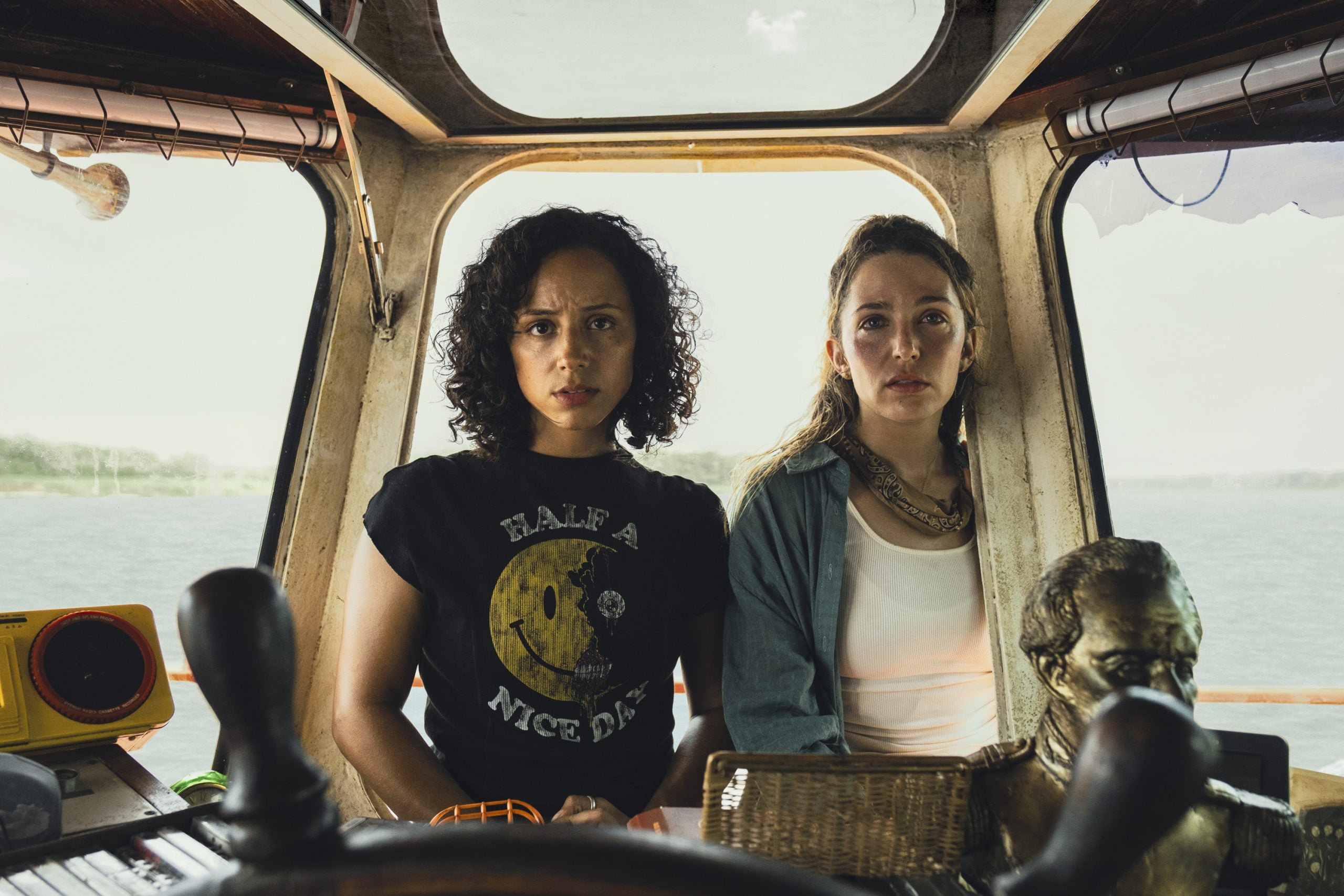Two women sit side by side in a boat cabin, looking forward with serious expressions. A yellow radio and a small statue are visible in the foreground.