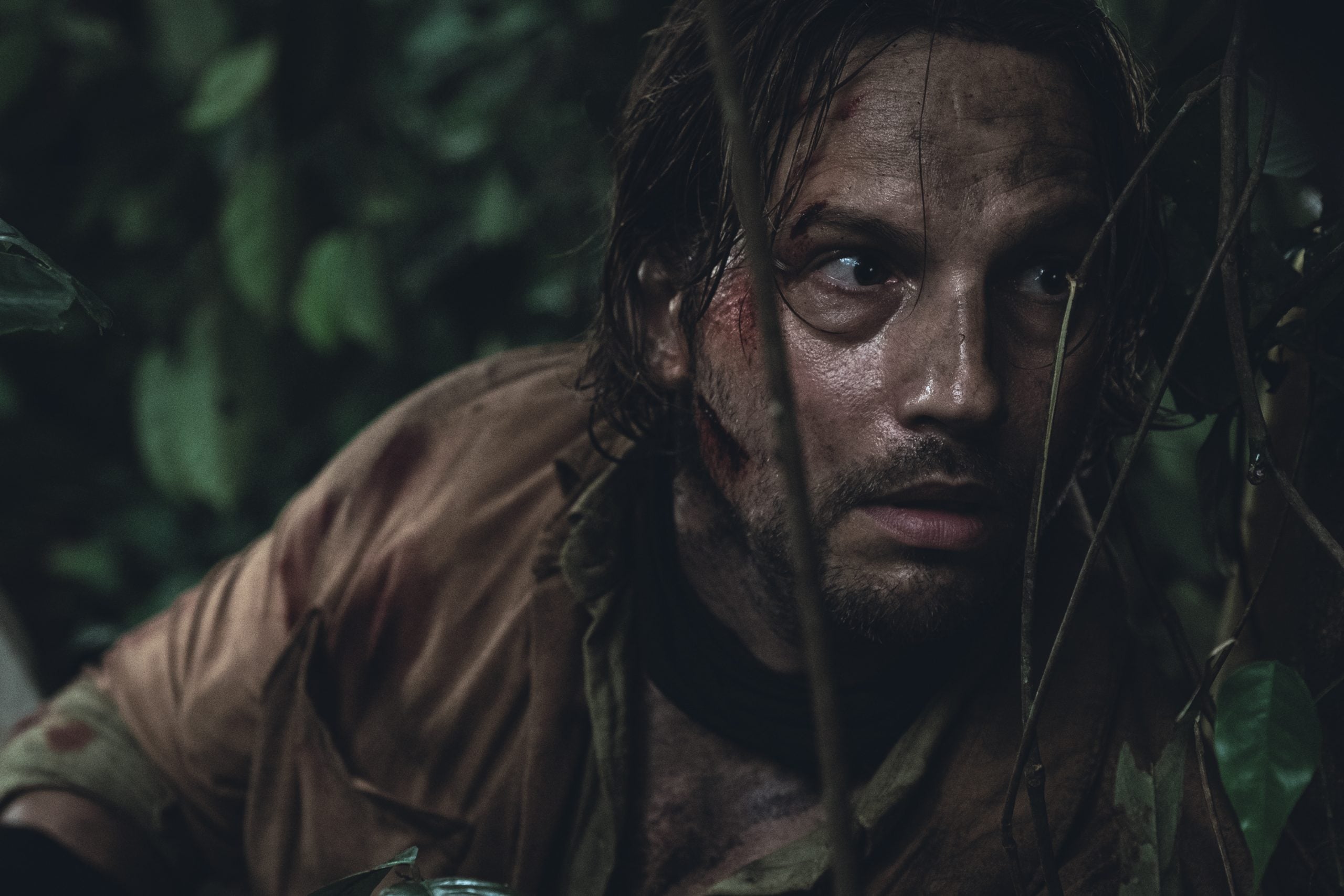 A man with a dirty, bruised face and disheveled hair crouches among jungle foliage, looking upward with a wary expression.